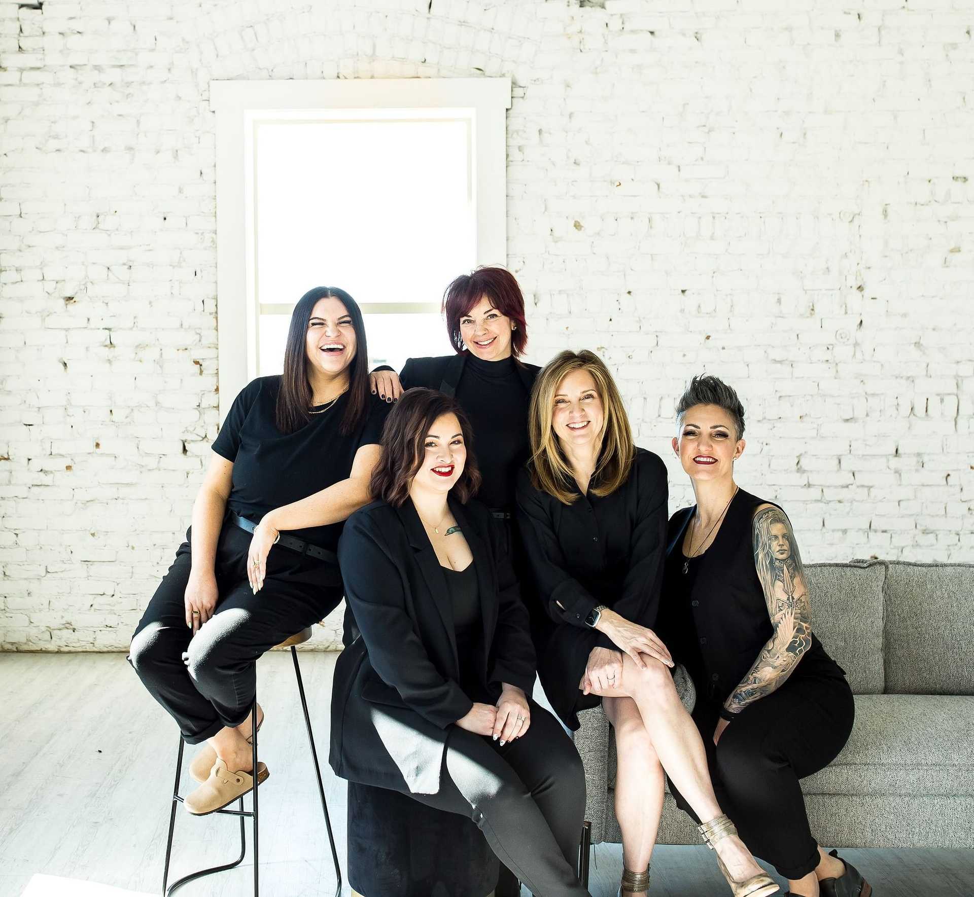 Five women in black smiling together in a white room.
